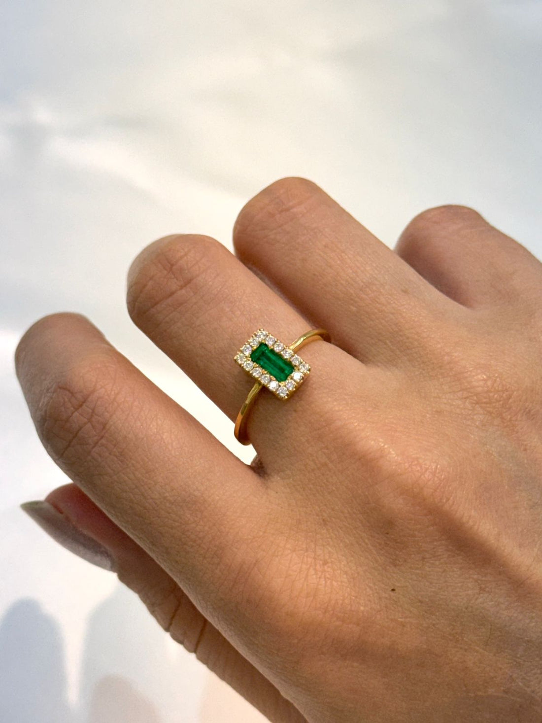 Emerald cut colombian emerald engagement ring in a diamond halo setting with a clean, polished 18K gold band being showcased on a hand with a neutral background