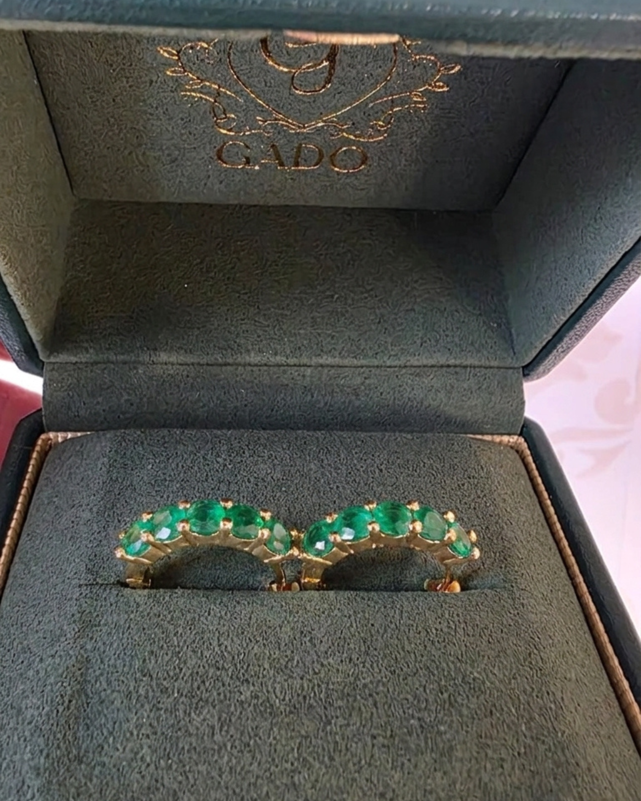 Gold ring with green gemstones in a jewelry box with 'GADO" branding.