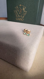 Gold earrings with green gemstones on a white cushion, with a green box labeled 'GADO' in the background.