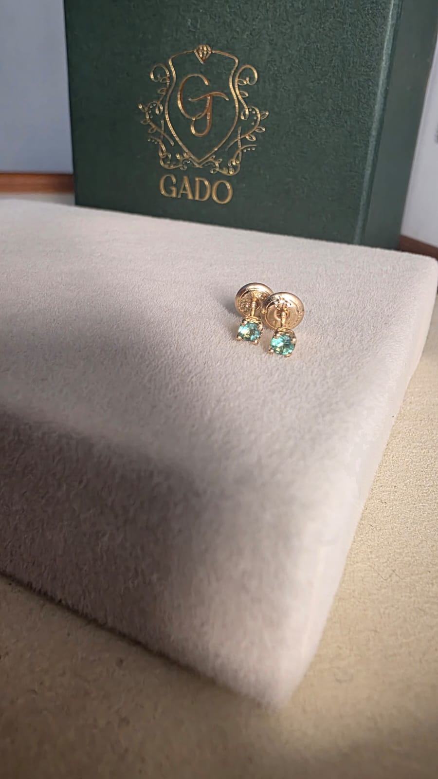 Gold earrings with green gemstones on a white cushion, with a green box labeled 'GADO' in the background.