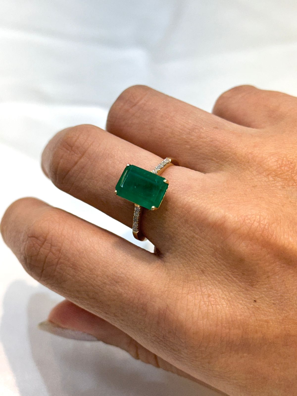Emerald cut Muzo Colombian Emerald ring, prong set,with partial pave diamond set band, 18K gold worn on a hand with a neutral background