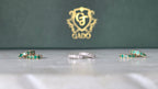 Jewelry set with emerald and diamond rings on a reflective surface, GADO logo in the background.