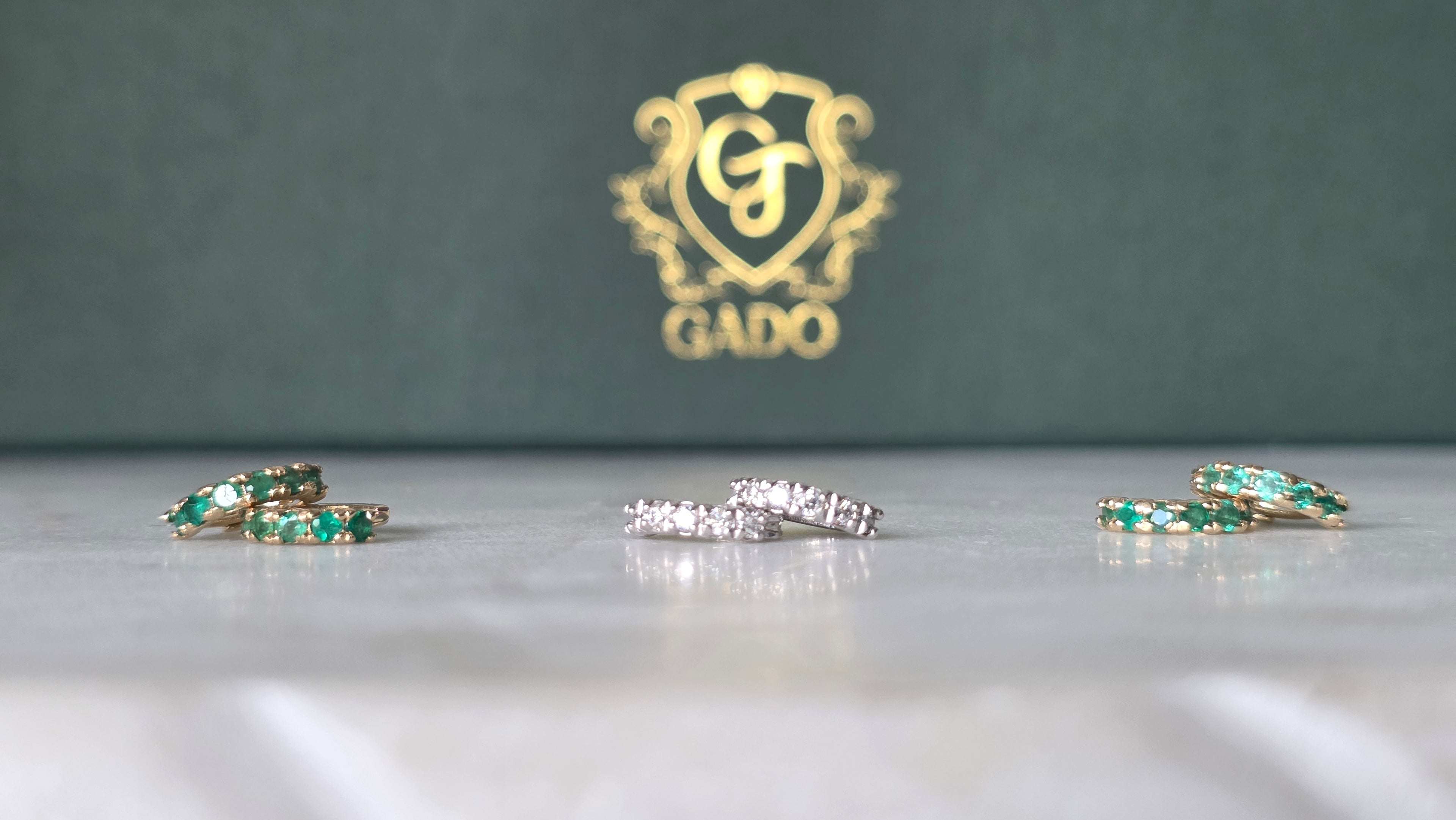 Jewelry set with emerald and diamond rings on a reflective surface, GADO logo in the background.