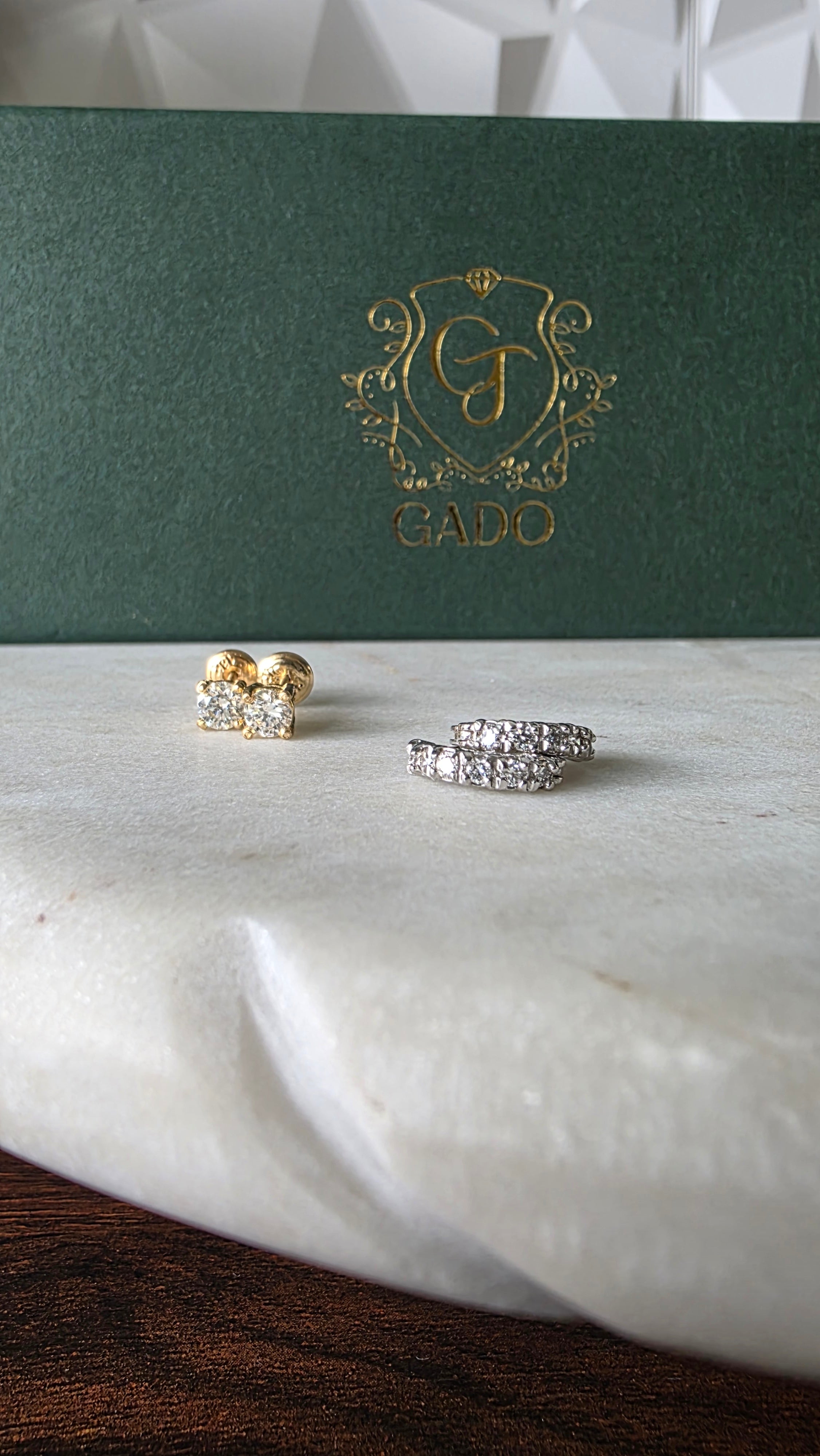Two diamond earrings on a white surface with a green jewelry box featuring a gold logo.